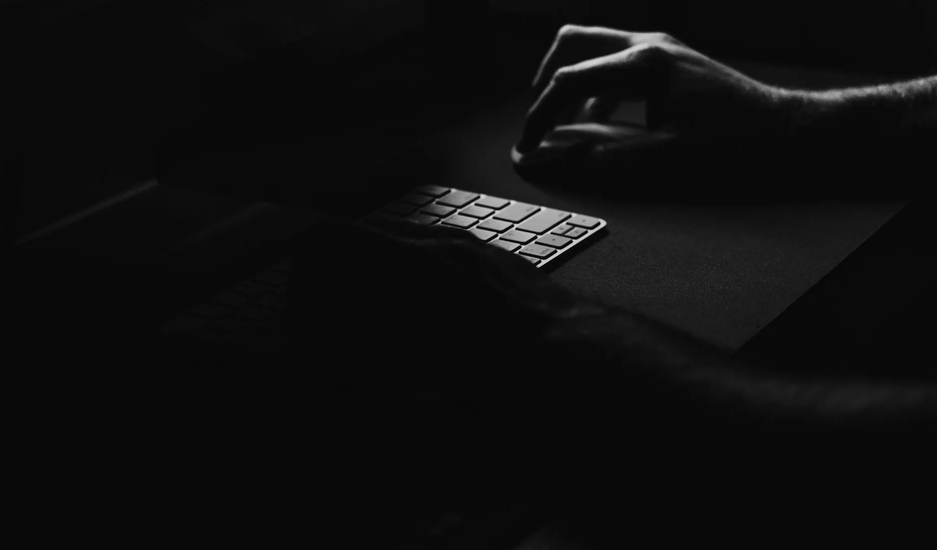 A hand on a mouse beside a keyboard lit by one screen in a dark room